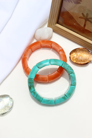 Peach Orbit Bangle Bracelets on a white surface with a wooden frame and seashells in the background