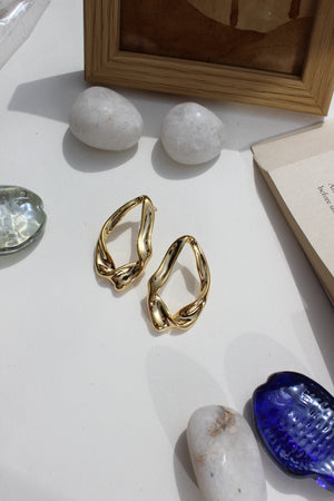 Golden Hug Earrings on a white surface with rocks and a blue seashell in the background
