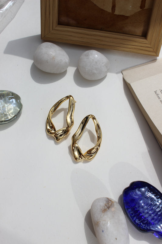 Golden Hug Earrings on a white surface with rocks and a blue seashell in the background