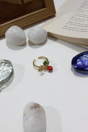 Sweetheart Spritz Ring with a red gemstone on a white surface next to stones and an open book