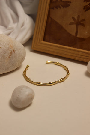Golden Reed Cuff Bracelet on a surface with stones and a framed picture in the background