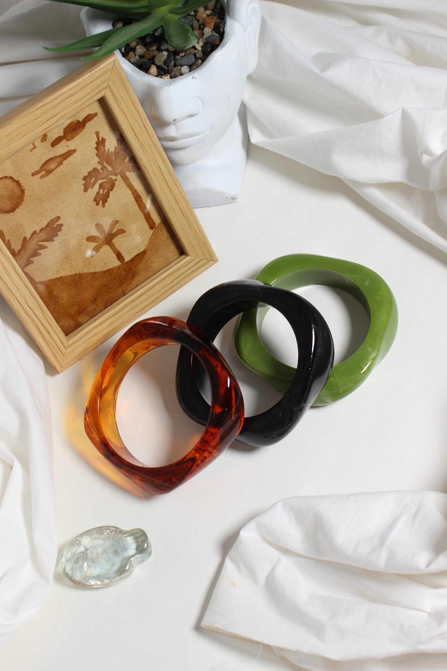 Aurora Stacks Resin Bangles on a white fabric background with a wooden frame and decorative stones