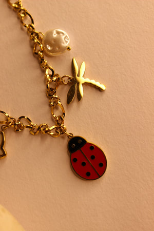 Bloom Folk Charms Necklace with various charms including a ladybug, dragonfly, and pearl on a pink background