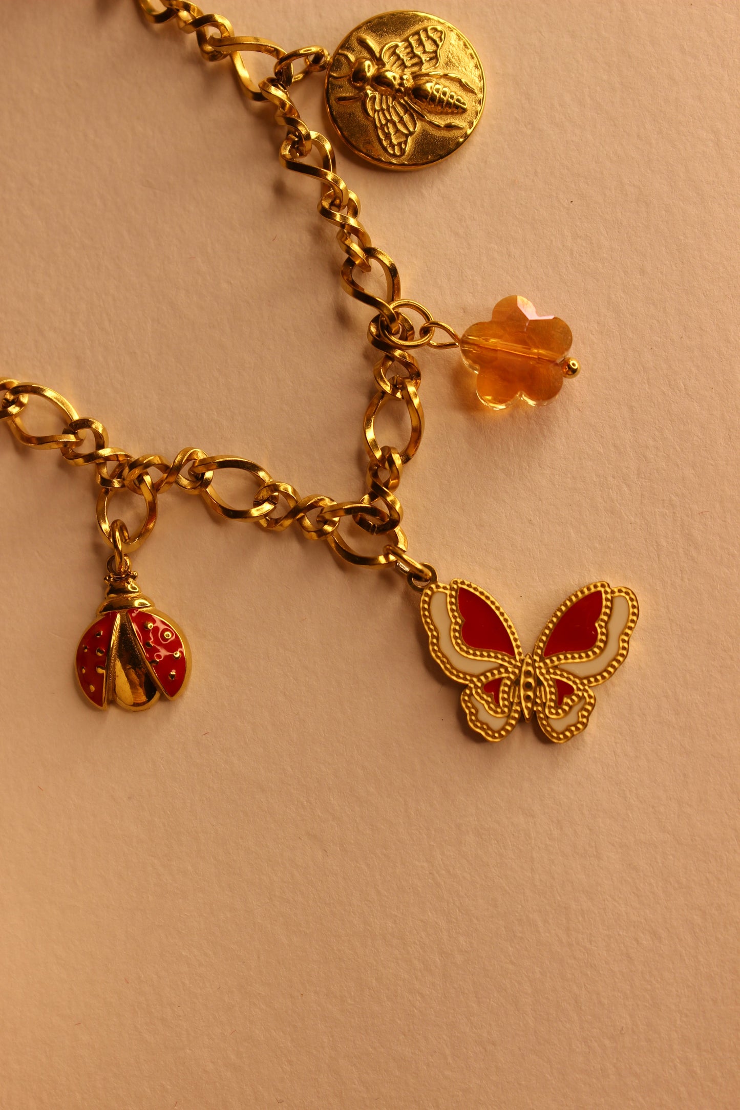 Gold charm necklace featuring butterfly, coin, and bead pendants displayed on white lace background