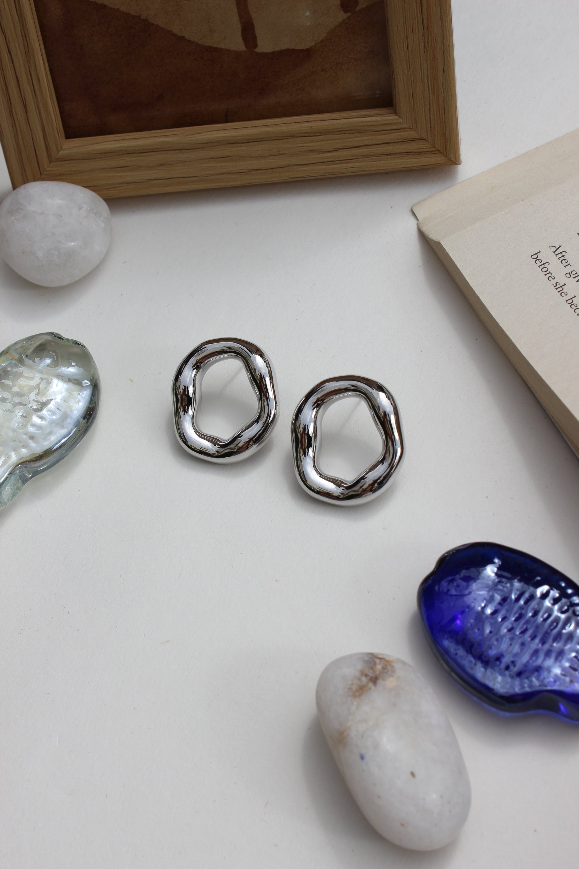 Lucid Frost Earrings on a white surface with a book, frame, and shell in the background