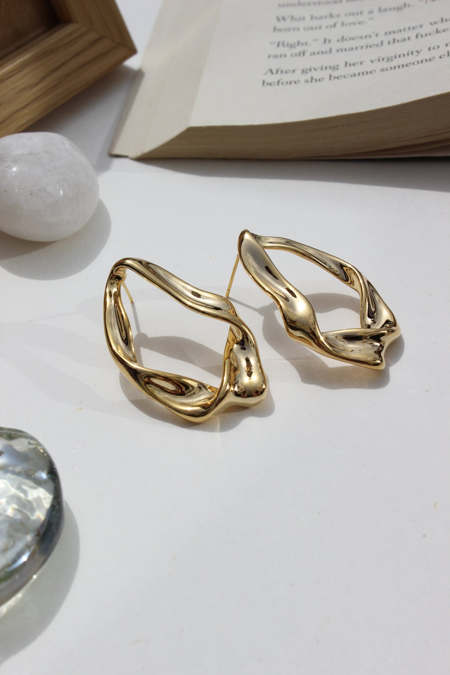 Golden Hug Earrings on a white surface with a blurred book and crystal in the background