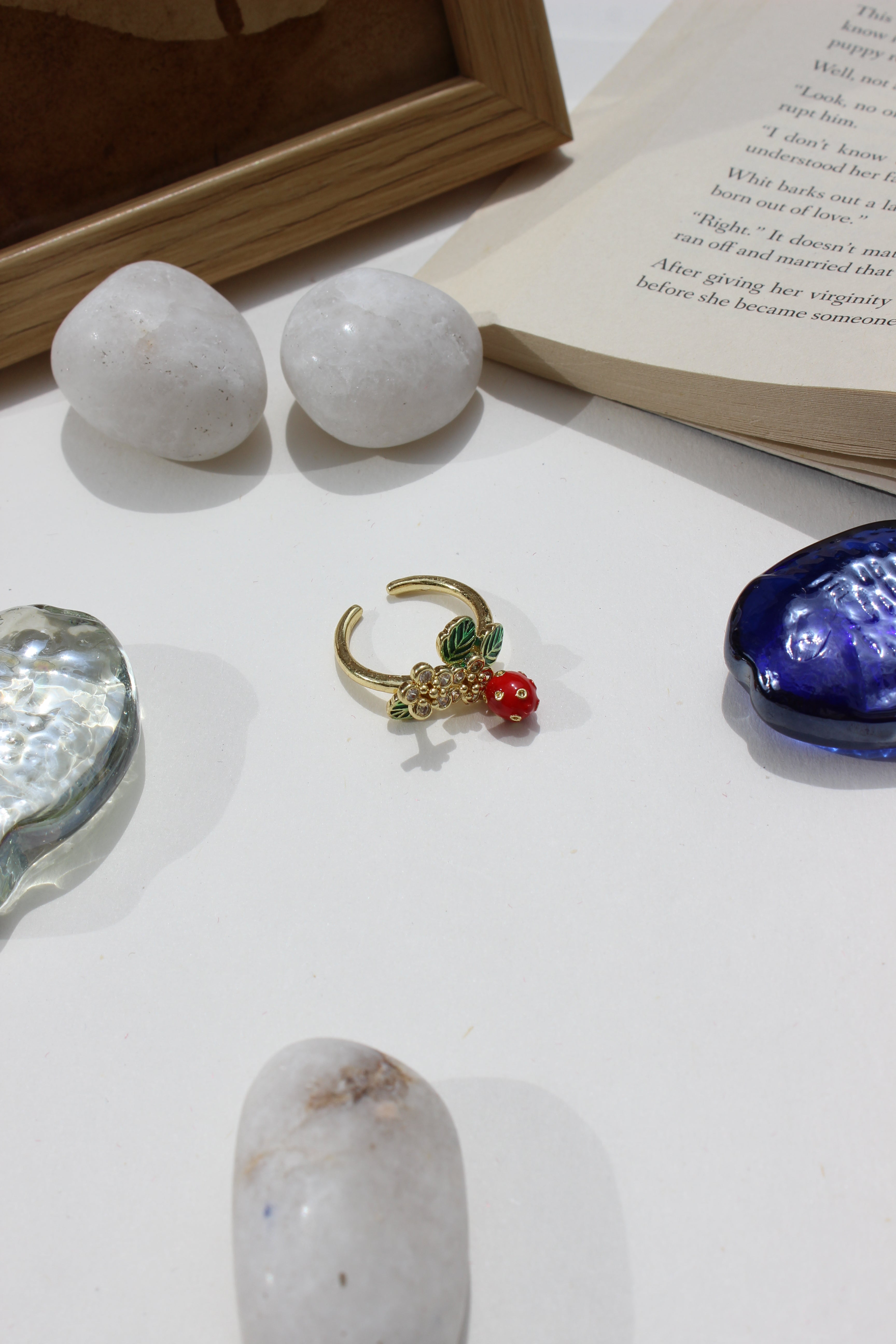 Sweetheart Spritz Ring with a red gemstone on a white surface next to stones and an open book