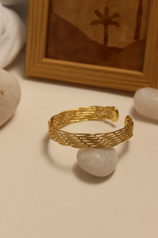 Woven Gold Cuff Bracelet on a beige surface with a wooden frame and stones in the background