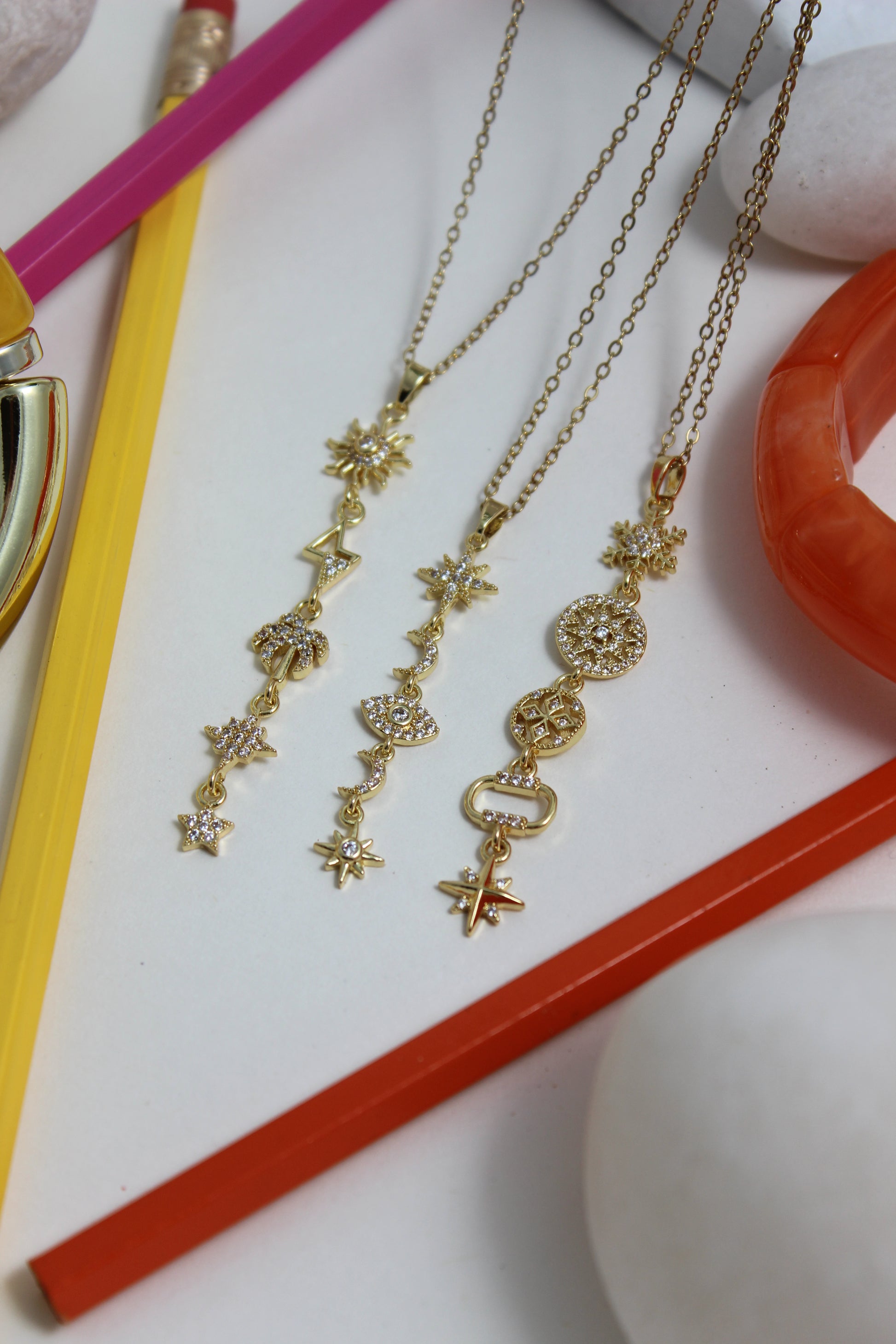 Golden Palm Necklace with star and bow designs on a white surface with red and yellow pencils
