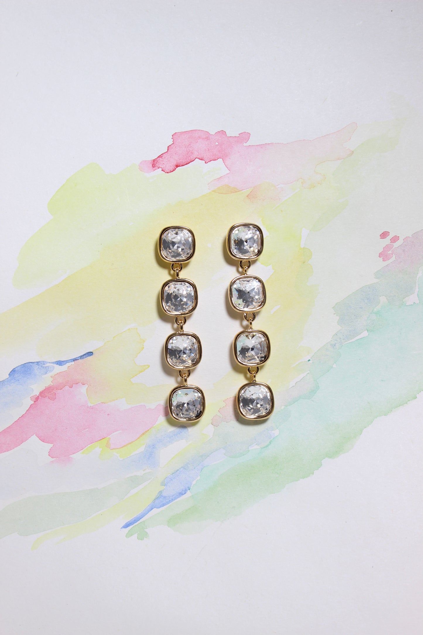 Pair of Glacier Glow Earrings with clear stones on a colorful watercolor background