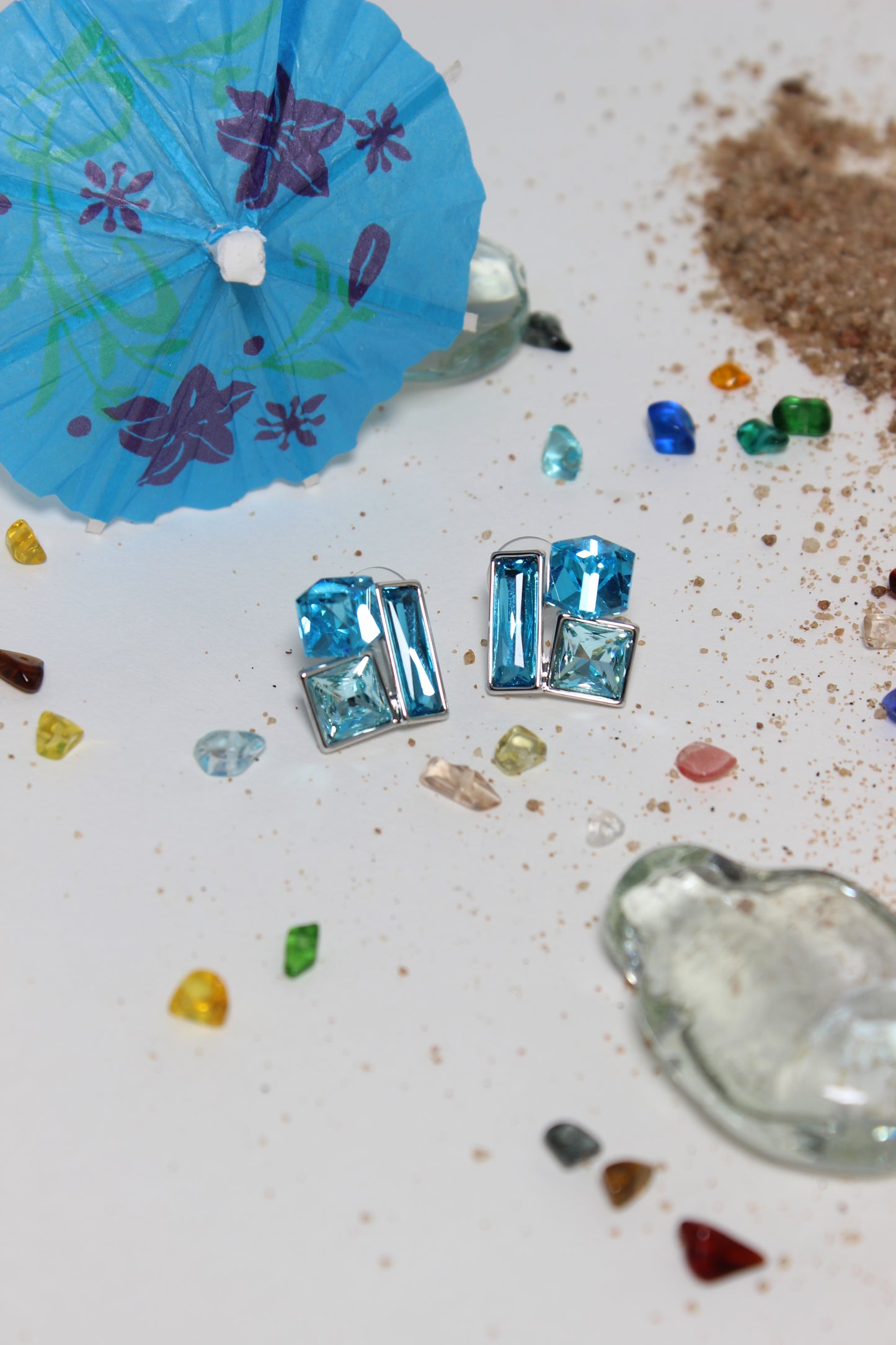 Blue gemstone Azure Glow Earrings on a textured surface with colorful stones and a blue paper umbrella