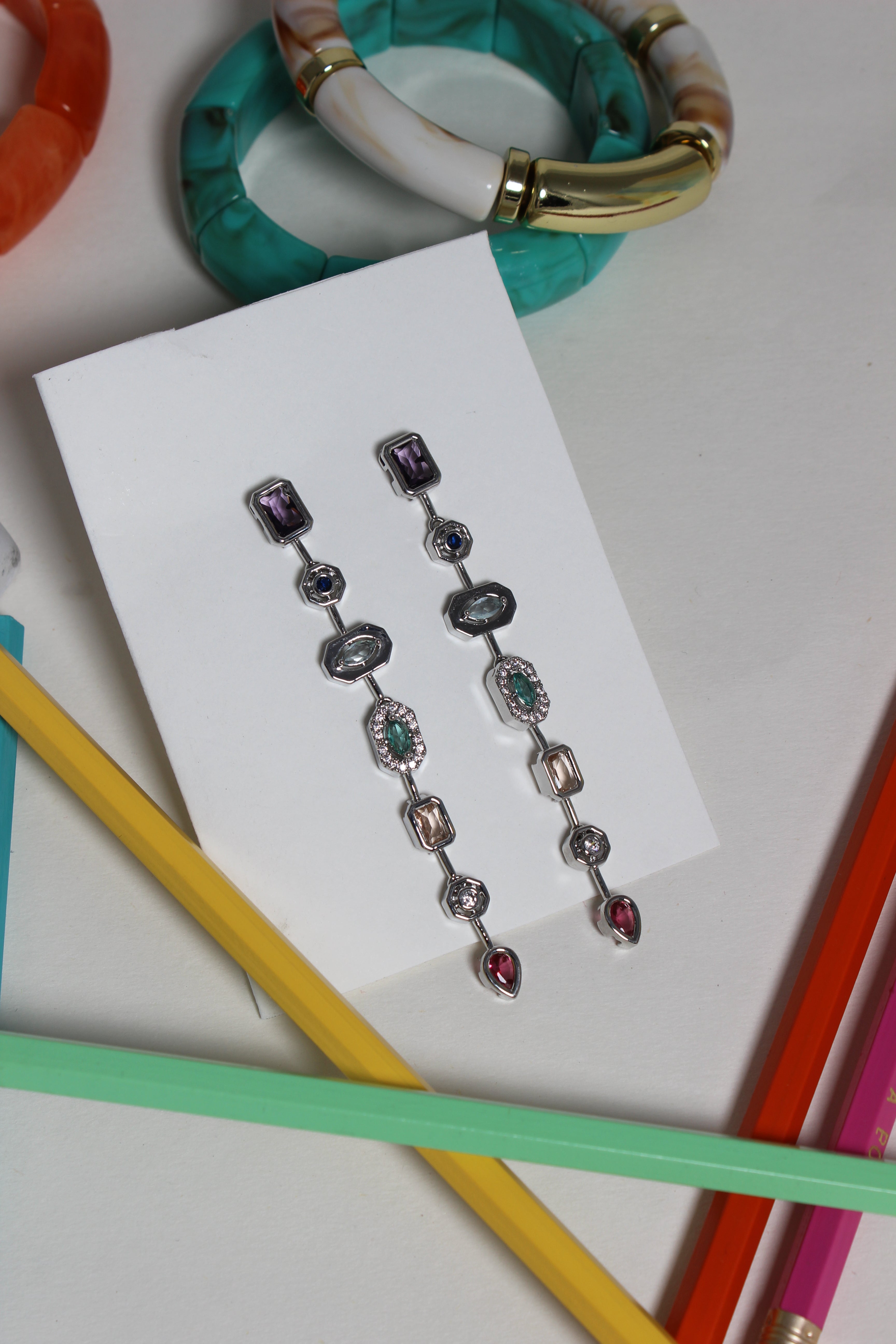 Argent Pastel Pour Earrings on a white card, surrounded by colorful pencils